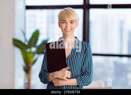 Entrepreneur Lady tenant dossier souriant debout près de la fenêtre dans le bureau Banque D'Images