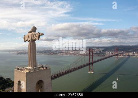 La statue du Christ Roi se dresse haute avec les bras tendus, surplombant le pont 25 de Abril et les eaux calmes du Tage à Lisbonne sous un ciel partiellement nuageux, vue aérienne Banque D'Images