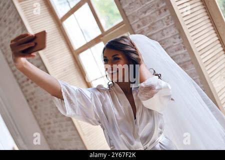 Charmante mariée. Jeune femme attrayante en peignoir de soie et voile souriant tout en prenant selfie près de la fenêtre Banque D'Images