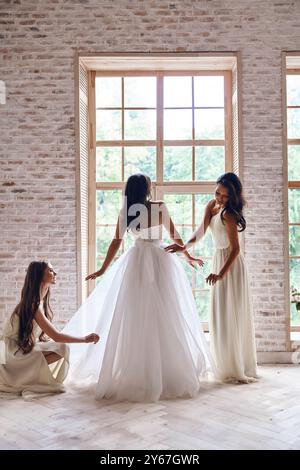 S'assurer qu'elle a l'air parfaite. Pleine longueur de demoiselles d'honneur aidant la mariée avec sa robe de mariée tout en se tenant près de la fenêtre ensemble Banque D'Images