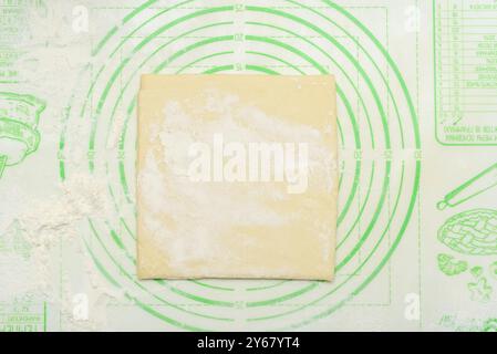 Un morceau carré de pâte est placé sur un tapis de pâte en silicone vert, légèrement saupoudré de farine. Le tapis comporte des repères de mesure et des diagrammes de cuisson. CON Banque D'Images