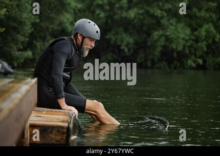 Portrait vue de côté d'un homme senior actif assis sur la jetée par ake avec les pieds dans l'eau et portant des équipements de protection pour l'espace de copie de sport Banque D'Images