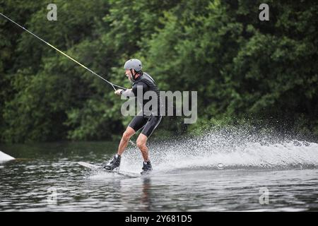 Plan d'action grand angle d'un homme âgé actif en train de faire du wakeboard sur un lac forestier pittoresque laissant un espace de copie de la piste des vagues Banque D'Images