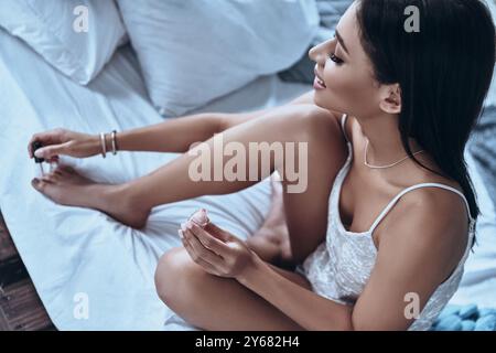 Tout devrait être parfait. Vue de dessus de jeune femme souriante attrayante appliquant du vernis à ongles aux orteils tout en étant assis sur le lit à la maison Banque D'Images