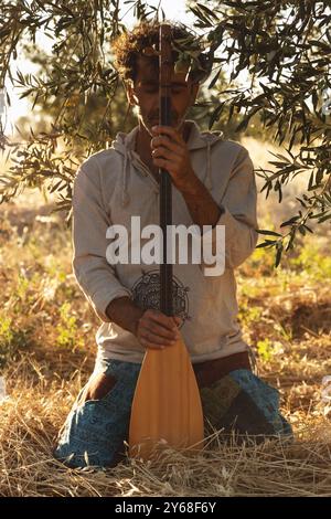 Un homme d'âge mûr est assis sur le sol dans un champ d'oliviers, tenant son baglama près, les yeux fermés dans le reflet. Le soleil filtre à travers l'olive br Banque D'Images