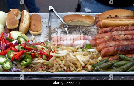 Vendeur de rue cuisant des hot dogs enveloppés de bacon, pinces tournant la nourriture. Cuisine populaire pour les foires et événements de rue Banque D'Images