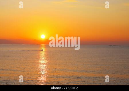 Un coucher de soleil orange vif se reflète sur la mer calme, avec un petit bateau passant à travers l'horizon alors que la journée touche à sa fin. Banque D'Images