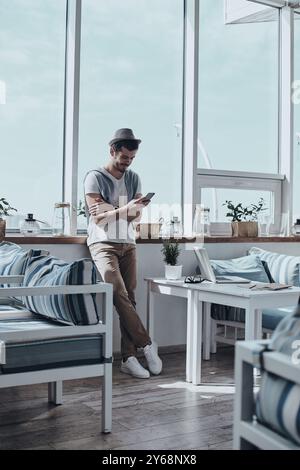 Vérification des messages des clients. Beau jeune homme tapant quelque chose à l'aide d'un téléphone intelligent tout en s'appuyant sur le rebord de la fenêtre dans le restaurant Banque D'Images