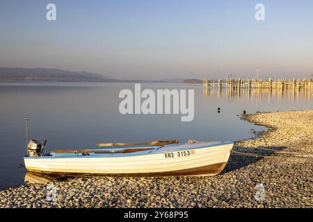 Un petit bateau blanc se trouve sur un lac tranquille avec une plage de galets à la lumière du matin, port, Allensbach, Bade-Wuerttemberg, Allemagne, Europe Banque D'Images