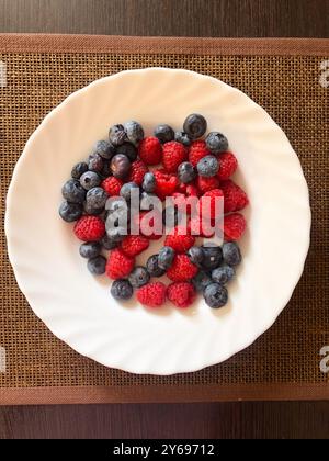 Vue de dessus d'une assiette blanche remplie de bleuets frais et de framboises, disposée sur un napperon tissé et une table en bois. Alimentation saine, végétalien biologique Banque D'Images