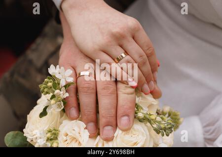 Gros plan romantique de mains entrelacées avec des anneaux de mariage et un bouquet de roses crème symbolisant l'amour et l'engagement. Banque D'Images