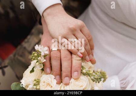 Gros plan romantique de mains entrelacées avec des anneaux de mariage tenant un bouquet de roses crème et de verdure. Banque D'Images