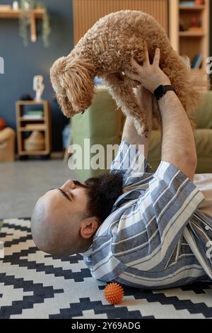 Homme avec la barbe complète soulevant chien aux cheveux bouclés tout en étant assis dans le salon conçu avec un décor moderne utilisant des étagères en bois et des plantes d'intérieur Banque D'Images