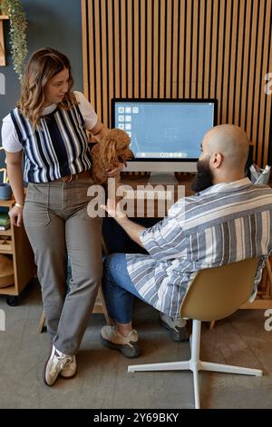 Femme et homme s'engageant avec un petit chien dans un espace de travail à la maison moderne avec un décor en bois créant une atmosphère chaleureuse et accueillante. Grand moniteur affichant l'arrière-plan du bureau Banque D'Images