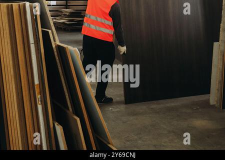 Comme la lumière du soleil filtre dans l'entrepôt, un travailleur diligent dans un gilet de sécurité lumineux transporte soigneusement une grande feuille de matériau au milieu de la stature imposante Banque D'Images
