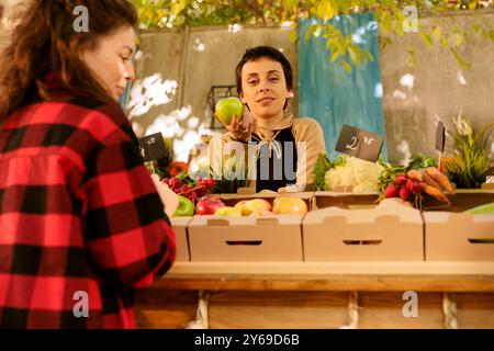 Joyeuse femme se tient derrière le stand du marché vendant des produits biologiques sains tout en tenant une pomme. La femelle du légumes regarde la caméra tout en exposant des fruits et légumes frais cultivés localement. Banque D'Images