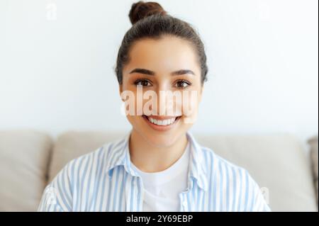 Photo en gros plan d'une magnifique jeune femme brune aux yeux bruns positive arabe, indienne ou caucasienne en chemise décontractée, assise sur un canapé dans le salon à la maison, regardant l'appareil photo, souriant amical Banque D'Images