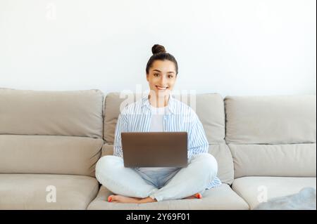 Confiant positive jolie femme brune indienne ou arabe, assise sur un canapé à la maison dans le salon, en utilisant son ordinateur portable pour le travail à distance sur un projet, sourit à la caméra. Photo de freelance féminine sur le canapé Banque D'Images