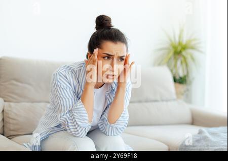 Bouleversée race mixte brune femme assise sur un canapé dans le salon après une dure journée de travail, souffrant de maux de tête, vertiges, migraine, besoin de traitement et de repos, masser les tempes. Stress, maux de tête, crampes Banque D'Images