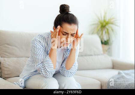 Stress, maux de tête, maladie. Femme de race mixte bouleversée assise sur un canapé dans le salon avec les yeux fermés, souffrant de maux de tête, vertiges, migraine, ayant des crampes dans un templess, besoin de traitement et de repos Banque D'Images