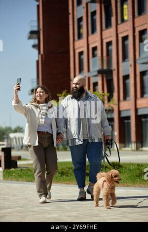 Couple heureux promenant chien tout en prenant selfie à l'extérieur près du complexe de construction moderne profitant d'une journée lumineuse et une atmosphère animée ensemble souriant tenant la main le long du chemin piétonnier Banque D'Images