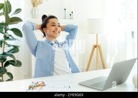 Heureux calme belle femme indienne ou arabe dans une chemise bleue, gestionnaire de bureau, assise sur un lieu de travail, se relaxant avec les mains derrière la tête, regardant loin, souriant, rêvant des week-ends Banque D'Images