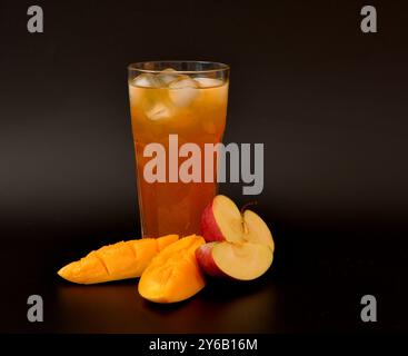 Jus de pomme-mangue dans un grand verre à facettes avec de la glace sur un fond noir, à côté de morceaux de fruits mûrs. Gros plan. Banque D'Images