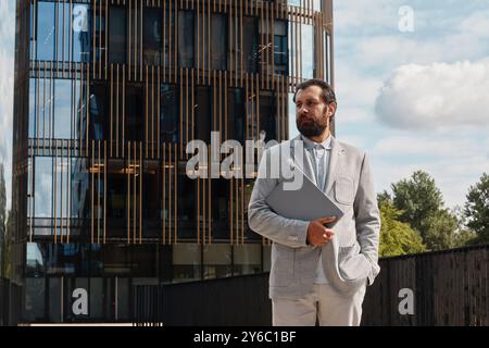 Homme d'affaires tenant la tablette tout en se tenant devant l'immeuble de bureaux moderne par une journée claire portant costume de couleur claire et regardant soigneusement dans la distance Banque D'Images