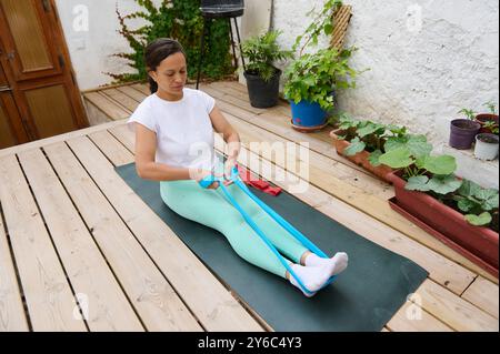 Une femme s'assoit sur un tapis de yoga dans une chemise blanche et des leggings turquoise légers, en utilisant une bande de résistance pour des exercices d'étirement sur une terrasse en bois avec du vert Banque D'Images