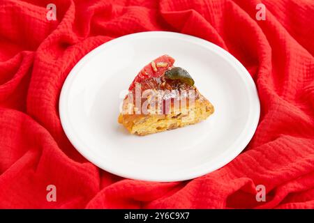 Une tranche de gâteau festif avec des fruits confits et des noix sur une assiette blanche, sertie sur un fond de tissu rouge Banque D'Images