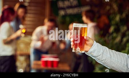 Bière à la main, prête pour la prochaine ronde de célébration. En arrière-plan, les fans se rassemblent autour de la table, levant leurs verres dans l'excitation et la camaraderie. Banque D'Images