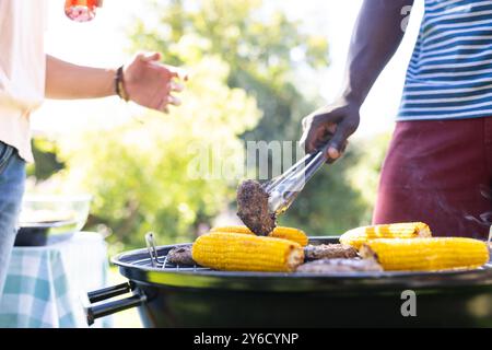 Griller du maïs et des hamburgers, deux amis masculins divers profitant d'un barbecue en plein air ensemble Banque D'Images