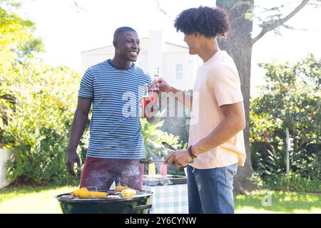 Griller du maïs sur barbecue, deux amis masculins divers appréciant des boissons et bavardant à l'extérieur Banque D'Images