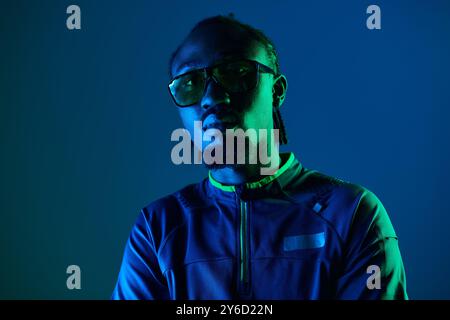 Un jeune homme en tenue tendance se tient confiant dans un studio de néon, éclairé par une lumière bleue frappante. Banque D'Images