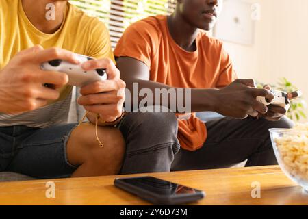 À la maison, jouer à des jeux vidéo, deux amis masculins divers utilisant des contrôleurs et assis sur le canapé Banque D'Images