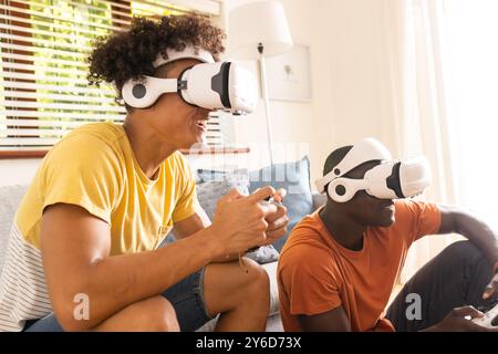 À la maison, jouer à des jeux vidéo, deux amis masculins divers utilisant des casques et des contrôleurs VR, appréciant t Banque D'Images