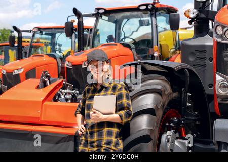 Revendeur de matériel agricole ou de construction. Femme en chemise à carreaux et casquette avec tablette numérique dans sa main appuyée sur la roue du nouveau tracteur. Banque D'Images