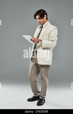 Un jeune et beau homme en tenue formelle examine une tablette tout en posant sur un fond gris élégant. Banque D'Images