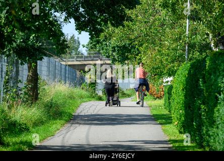 Deux personnes profitent d'une journée ensoleillée dans un parc ; une femme se promène avec une poussette tandis qu'une autre fait du vélo le long d'un chemin bordé d'arbres près d'une clôture Banque D'Images