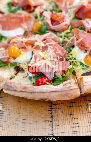 Gros plan de pizza artisanale garnie de prosciutto, roquette fraîche, tomates cerises jaunes et rouges colorées et fromage râpé. Servi sur bois rustique Banque D'Images