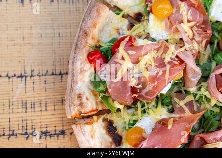 Gros plan de pizza artisanale garnie de prosciutto, roquette fraîche, tomates cerises jaunes et rouges colorées et fromage râpé. Servi sur bois rustique Banque D'Images