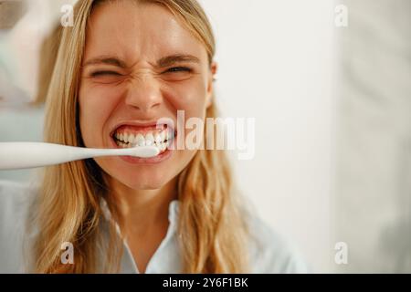 Une jeune femme se brossant joyeusement les dents avec une brosse à dents électrique dans sa salle de bain Banque D'Images