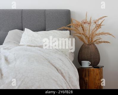 Vase en céramique marron avec herbe sèche, tasse de café blanche avec soucoupe et un téléphone portable couché sur une table de chevet tronc d'arbre. Banque D'Images