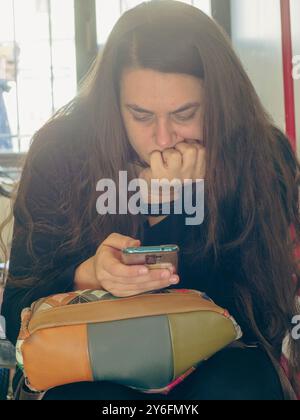 Une femme inquiète est assise à l'intérieur près d'une fenêtre, se mord nerveusement les ongles tout en lisant un texte sur son smartphone Banque D'Images