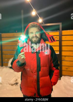 L'homme barbu dans une veste rouge tient un étincelant brillant, debout à l'extérieur par une nuit d'hiver enneigée, avec des guirlandes lumineuses festives suspendues au-dessus de la tête. Noël Banque D'Images