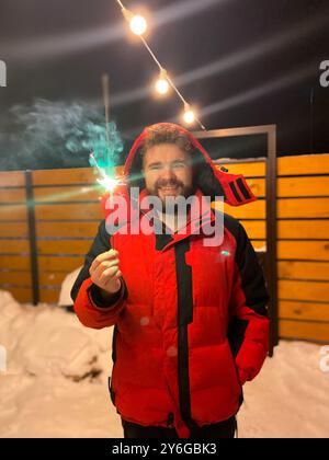 L'homme barbu dans une veste rouge tient un étincelant brillant, debout à l'extérieur par une nuit d'hiver enneigée, avec des guirlandes lumineuses festives suspendues au-dessus de la tête. Noël Banque D'Images