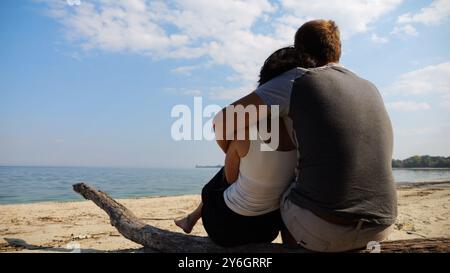 Couple romantique embrasse pendant assis sur le rondin de bois sur la plage. Paire heureuse admirant une mer et un paysage. Amoureux passant du temps ensemble à la station Banque D'Images