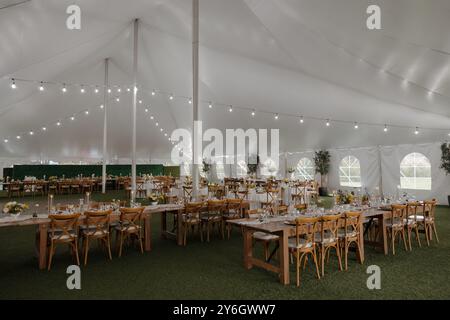 Configuration élégante de réception de mariage à l'intérieur d'une grande tente blanche avec guirlande lumineuse et tables et chaises en bois. Banque D'Images