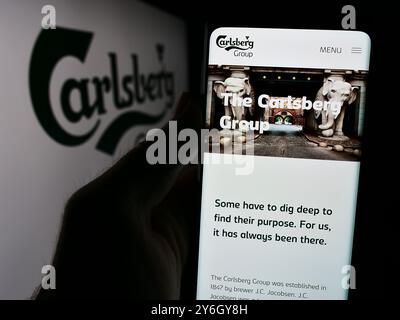 Dans cette illustration photo, une personne tient un téléphone portable avec la page Web de la brasserie danoise Carlsberg COMME devant le logo de l'entreprise. Banque D'Images