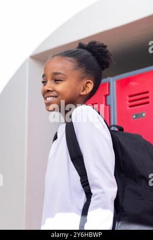 Fille afro-américaine souriante avec sac à dos debout près des casiers de l'école, prête pour la classe Banque D'Images